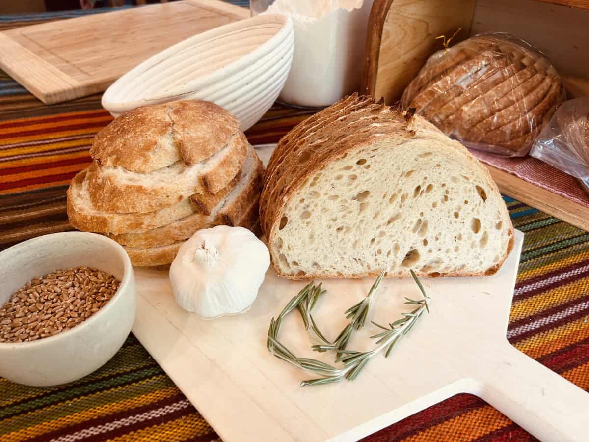 Whole and sliced loaves of bread on a breadboard with a head of garlic and herbs